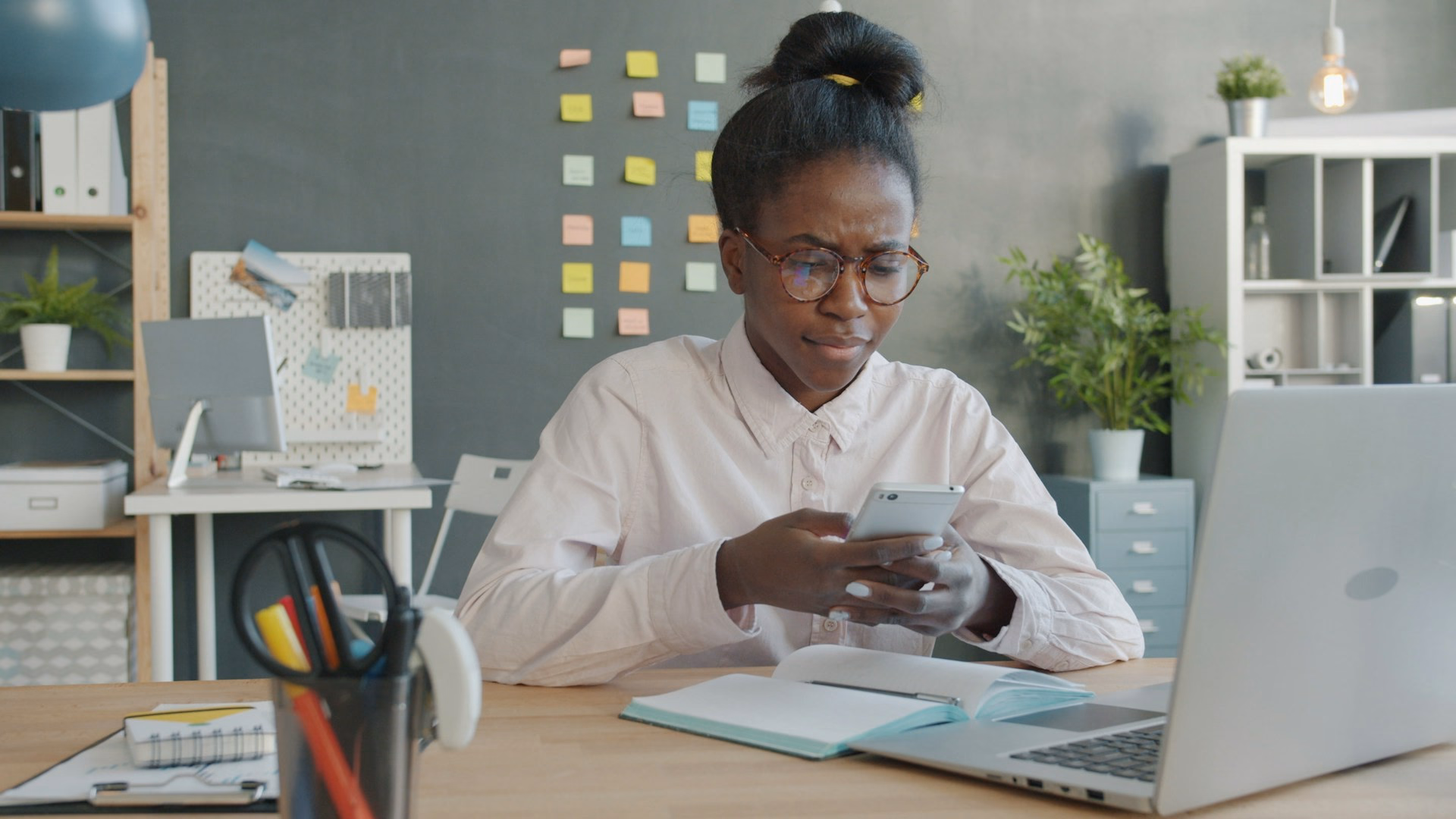 Eine Frau sitzt am Schreibtisch im Büro, schaut konzentriert auf ihr Smartphone, daneben ein aufgeklappter Laptop und ein Notizbuch.