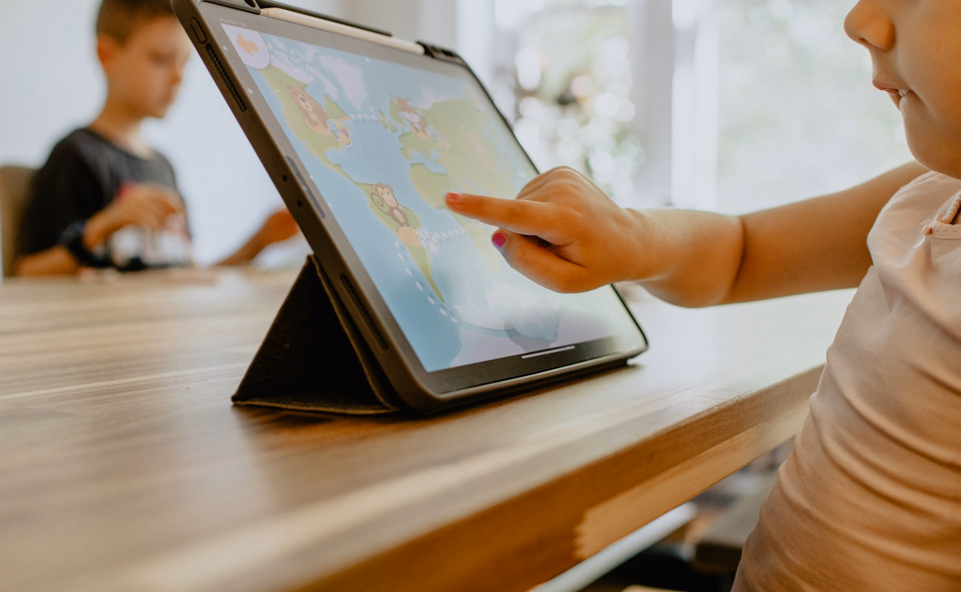 Kind tippt am Tisch auf einem Tablet, das in einer Hülle aufgestellt ist.