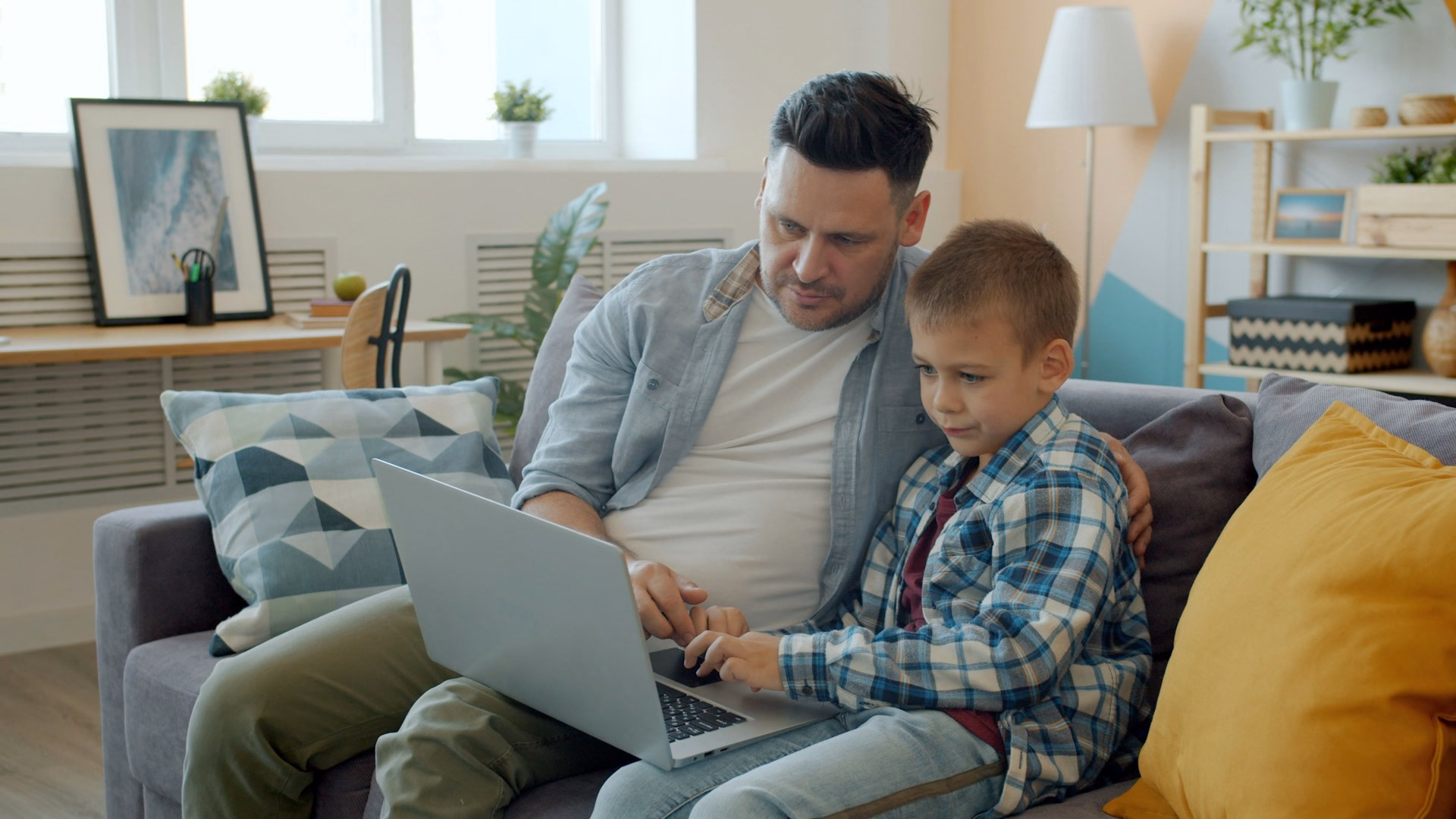 Vater und Sohn sitzen gemeinsam mit einem Notebook auf dem Sofa.