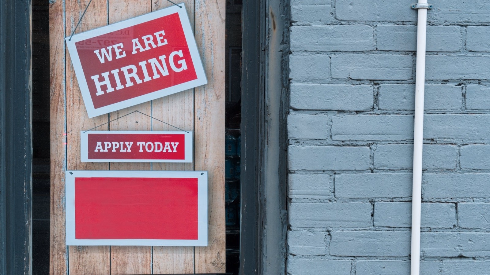 Rote Schilder „We are hiring“ und „Apply today“ hängen in einem Schaufenster neben einer hellgrauen Backsteinwand.