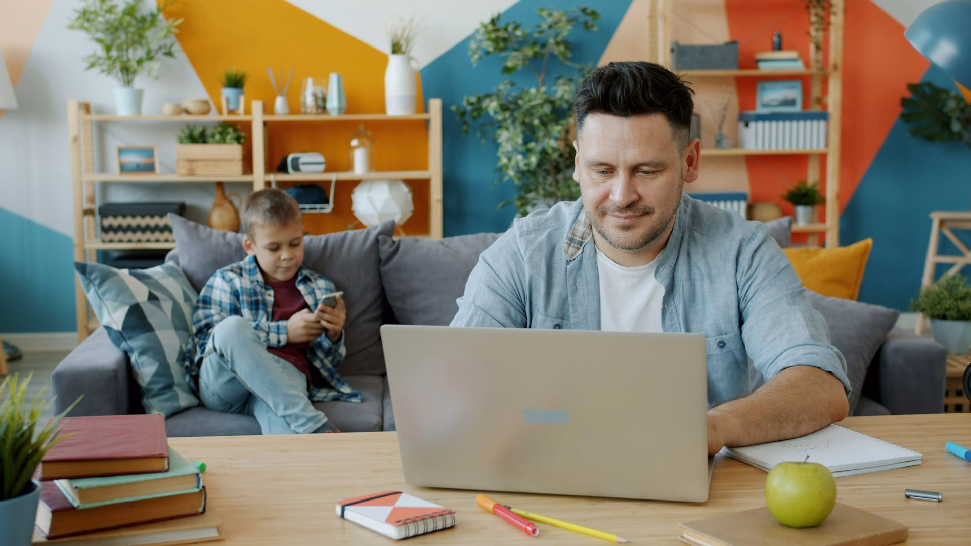Vater arbeitet im Wohnzimmer am Notebook, im Hintergrund sitzt sein Sohn mit einem Smartphone auf dem Sofa – Technik im deutschen Haushalt.