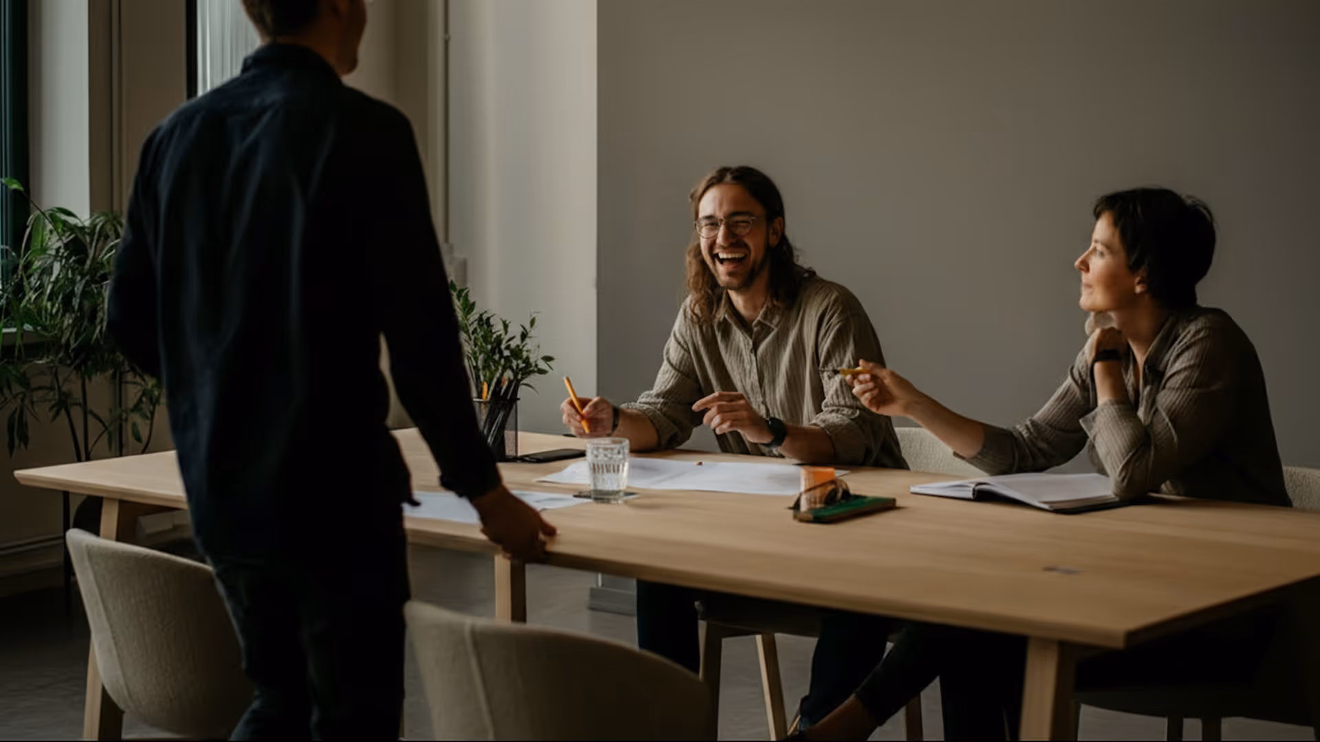 Zwei Personen sitzen an einem Holztisch und unterhalten sich mit einem stehenden Mann in einem modernen Büro.