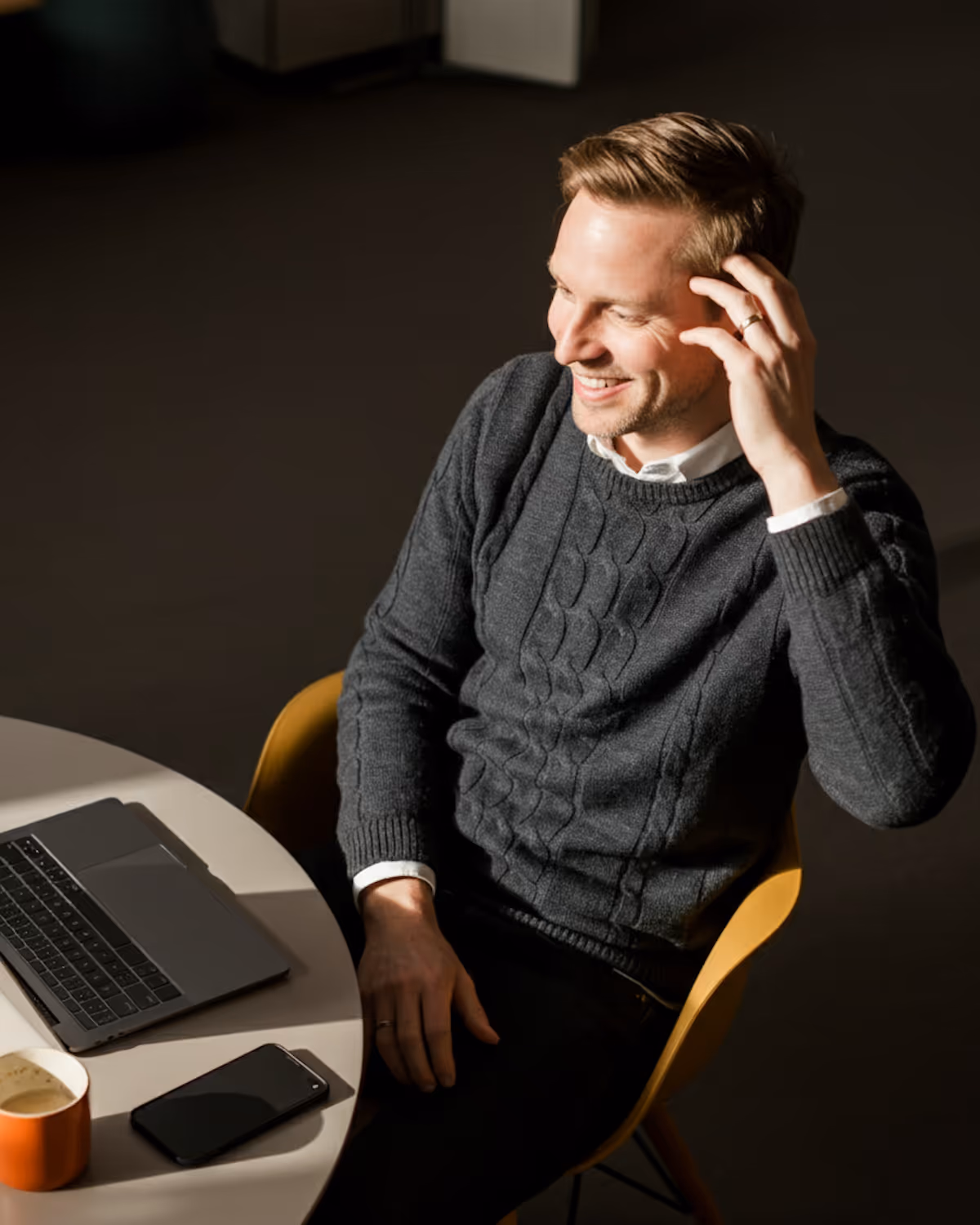 Mann mit grauem Pullover sitzt an Tisch mit Laptop, Smartphone und Tasse, lächelt und berührt sein Haar.